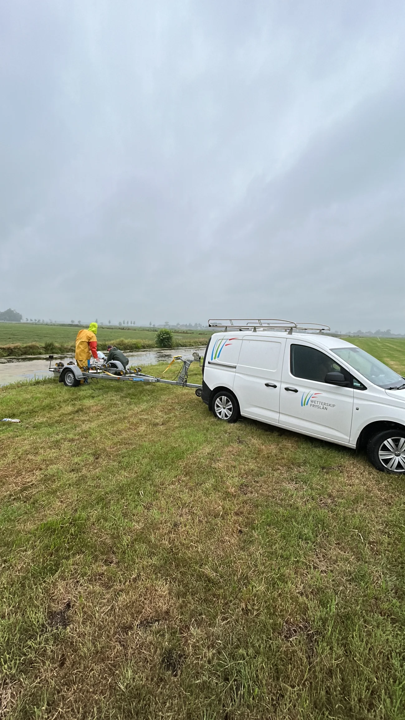 Een witte bestelbus met het logo van Wetterskip Fryslân staat in het weiland naast een poldersloot, met een boottrailer erachter.
